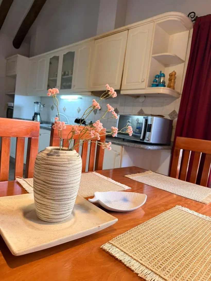 La mesa del comedor con individuales y un florero, y la cocina de la casa detrás
